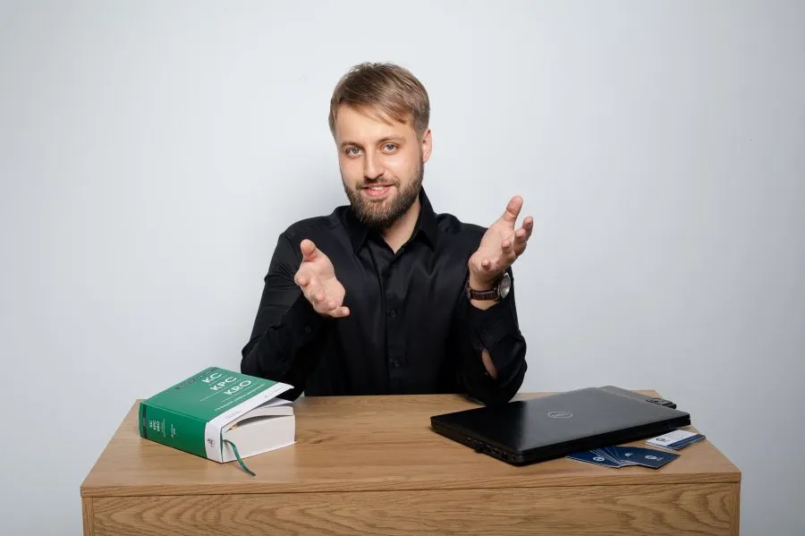Zdjęcie przedstawia prawnika siedzącego przy biurku, na którym znajdują się kodeks prawny, laptop oraz dokumenty. Mężczyzna wykonuje otwarty gest dłońmi, sugerujący rozmowę lub wyjaśnianie zagadnień prawnych. Fotografia ilustruje temat prawa gospodarczego i obsługi firm, podkreślając profesjonalne doradztwo prawne dla przedsiębiorców w zakresie prowadzenia działalności oraz bieżącego wsparcia biznesu.
