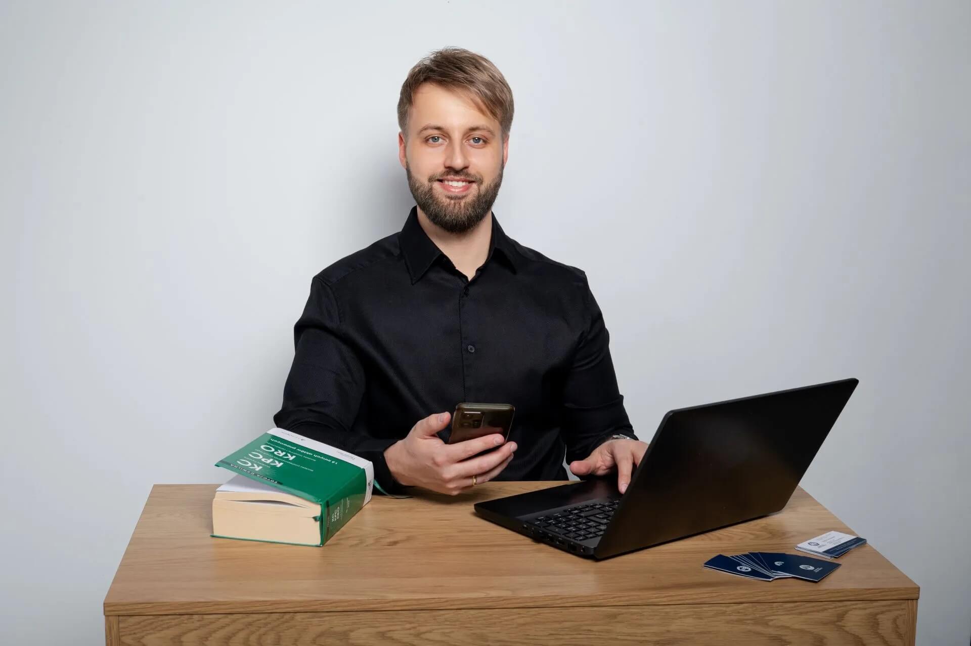 Na zdjęciu widać mężczyznę siedzącego przy drewnianym biurku, ubranego w czarną koszulę. Przed nim znajduje się otwarty laptop, a w dłoni trzyma smartfon. Na biurku leży również książka dotycząca Kodeksu cywilnego (KC) oraz kilka wizytówek. Mężczyzna patrzy w stronę aparatu i się uśmiecha. Jasne, neutralne tło oraz profesjonalna aranżacja zdjęcia nadają mu biznesowy i prawniczy charakter.