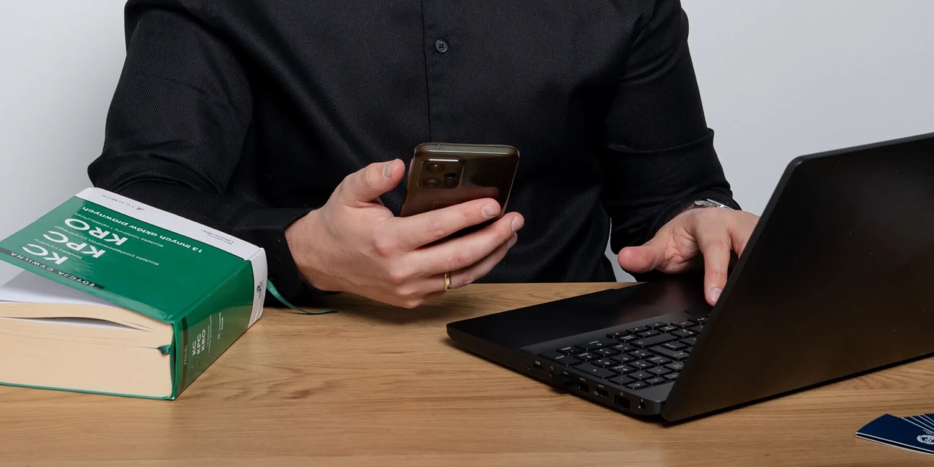 Na zdjęciu widać mężczyznę pracującego przy biurku, korzystającego jednocześnie ze smartfona i laptopa. Obok znajdują się książki prawnicze, co podkreśla profesjonalny i związany z prawem charakter fotografii.